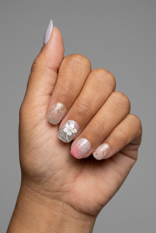 A close-up of a hand showing off a set of decorated press-on nails. The nails feature a mix of glittery pink, sheer nude, and white floral designs with gold foil accents.
