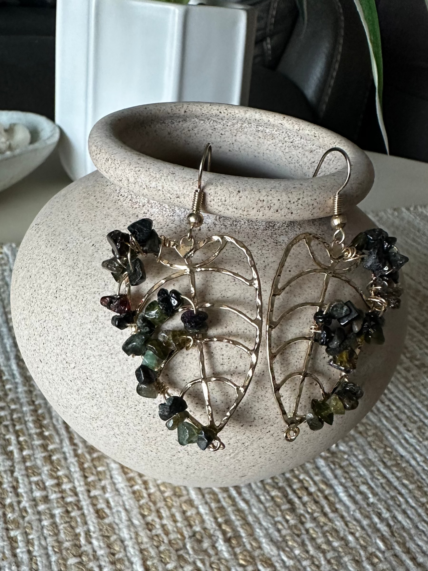 Monstera leaf shaped earrings with green semi-precious gemstones on a textured surface.