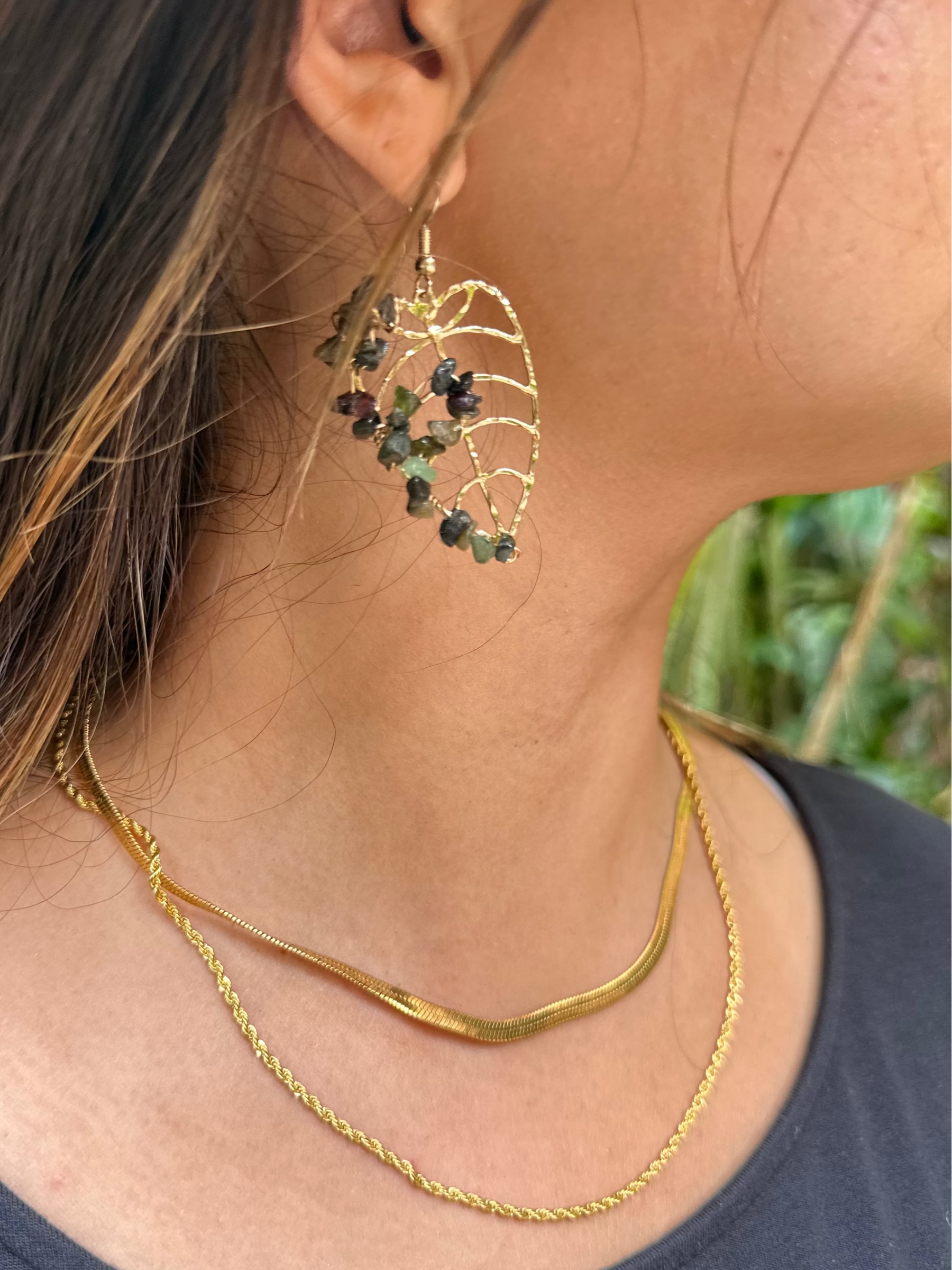 Close-up of a young woman wearing monstera earrings with green semi-precious gemstones and layered rope and herringbone necklace.
